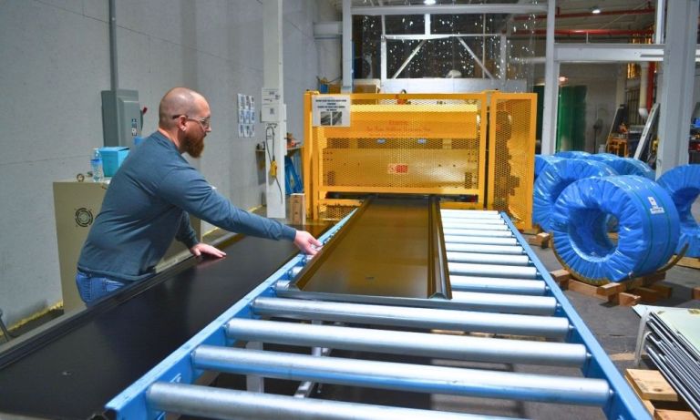 Worker guiding a metal roof panel along a blue conveyor machine.