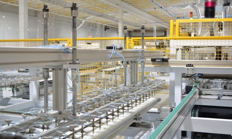 A close-up view of automated robotic machinery with vertical shafts and pneumatic tubes along a factory production track enclosed by yellow safety fencing.
